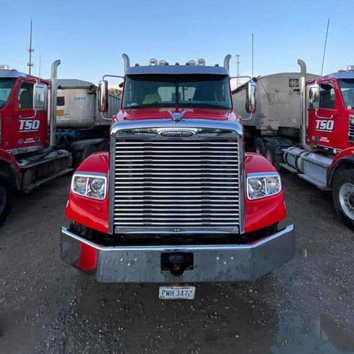 Front view of a red Freightliner dump-truck tractor with a chrome grille and bumper between two other red trucks.