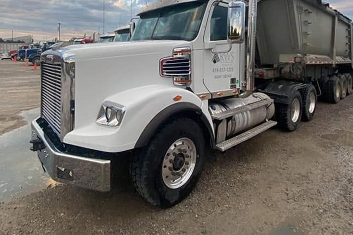 White tractor with an aluminum dump trailer under a cloudy dusk sky.