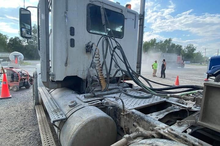 Hoses and cables across the back of a dump-truck cab, with two workers far off on a gravel yard.