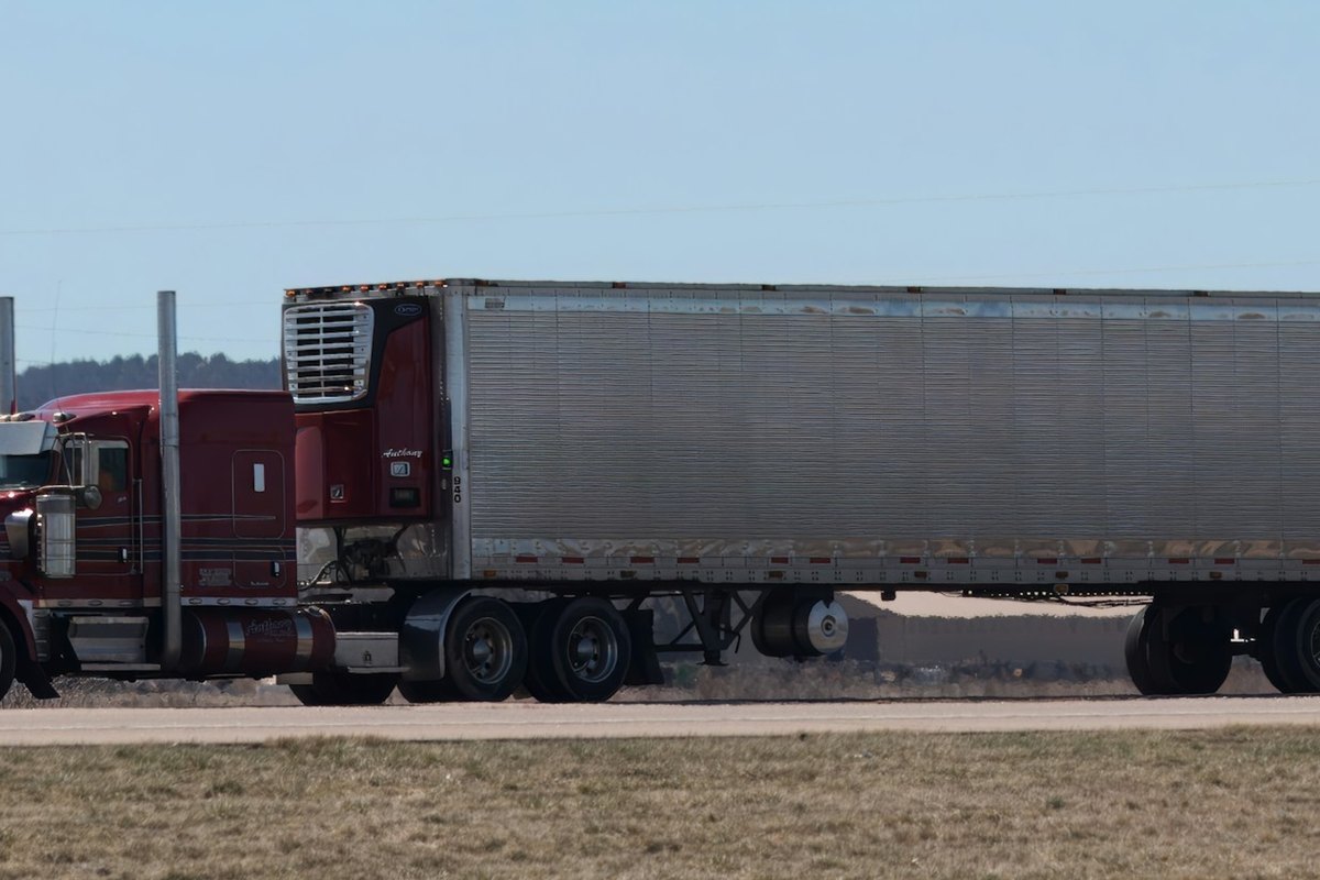 Ribbed silver refrigerated trailer with its refrigeration unit behind a dark red cab on a highway.