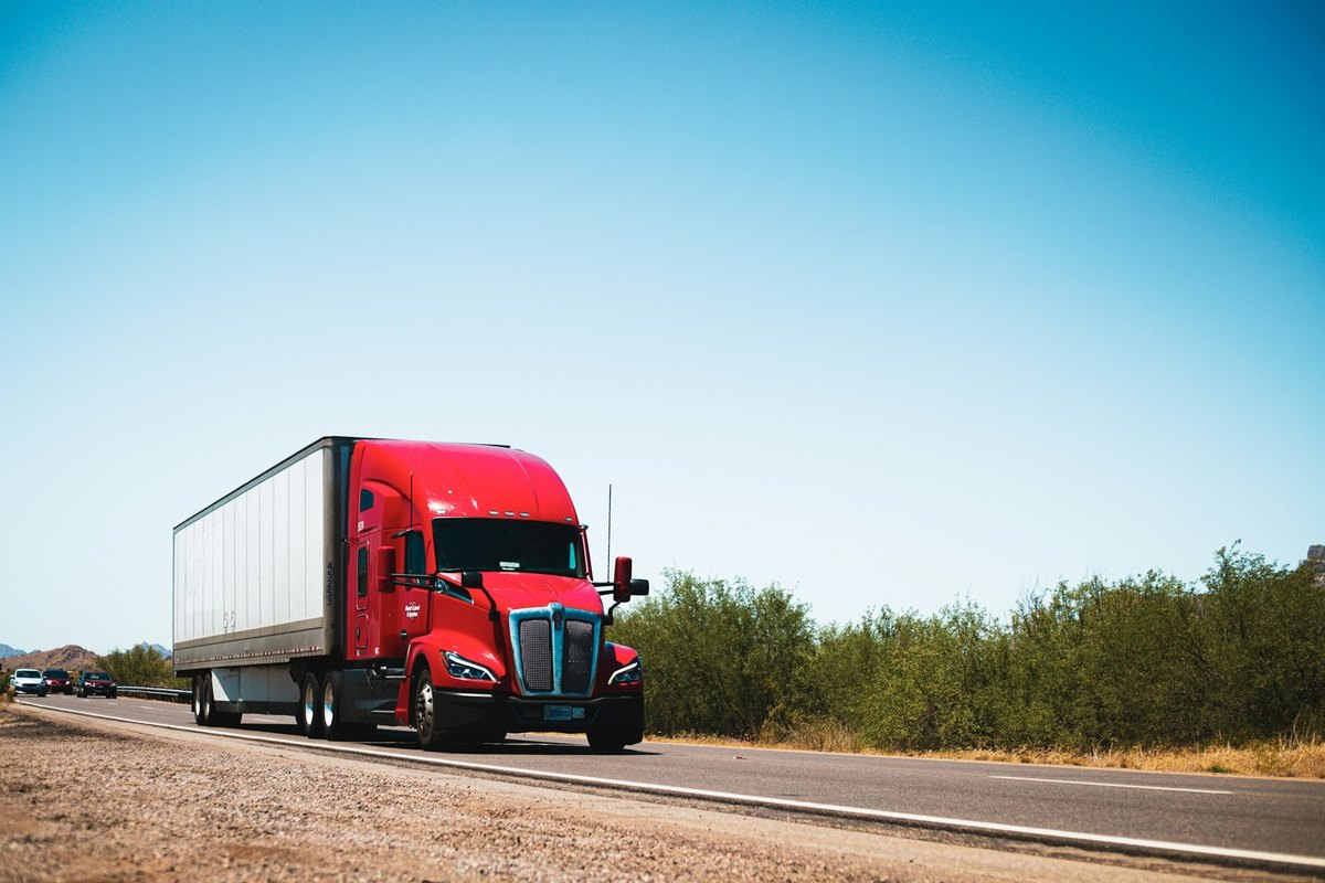 Red tractor with a white dry box trailer driving toward the camera on a two lane highway under a clear sky.