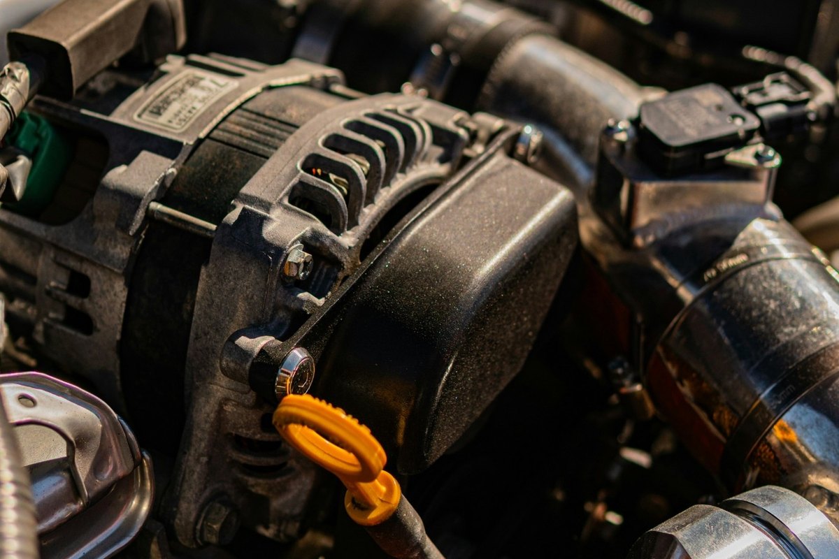 Close-up of a dusty engine bay with an alternator, an orange dipstick handle, hoses and a metal strut bar.