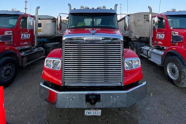 Front of a red Freightliner dump-truck tractor with a chrome grille and bumper between two other red trucks.