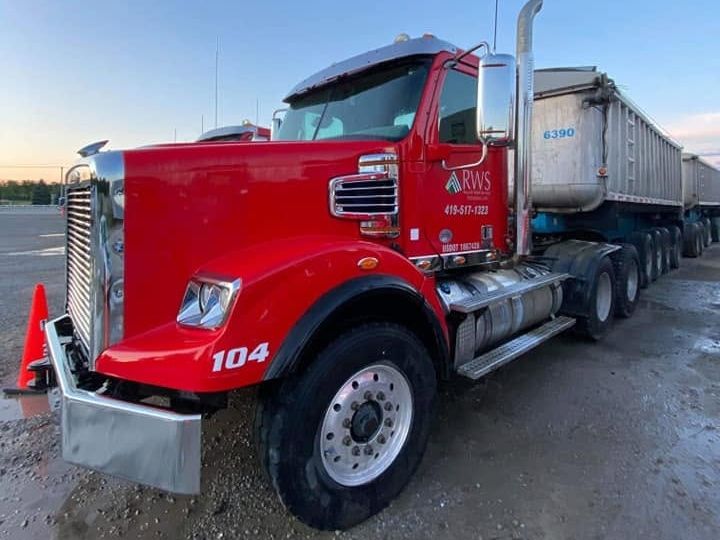 Cab of a red tractor with a chrome mirror and stack, one aluminum trailer at its side, clear dusk sky.