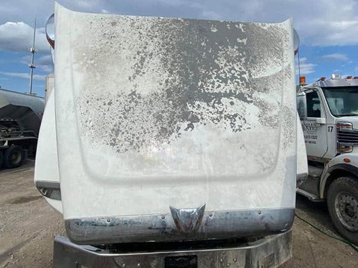 Front of a white truck with the hood raised and gray road film and mineral spots on the hood top.
