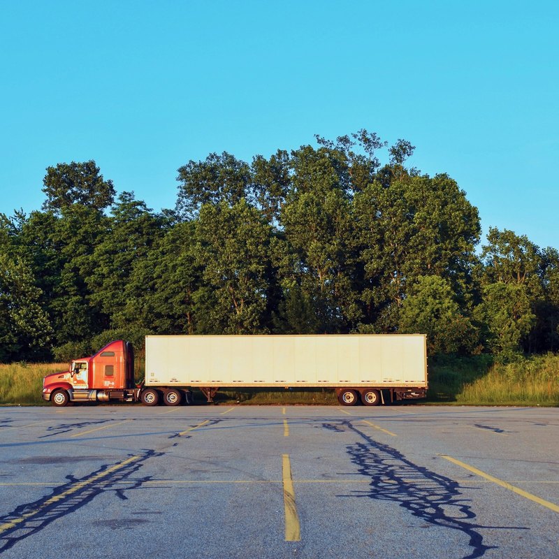 Red tractor and a plain white trailer parked across an empty asphalt lot in front of broadleaf trees.
