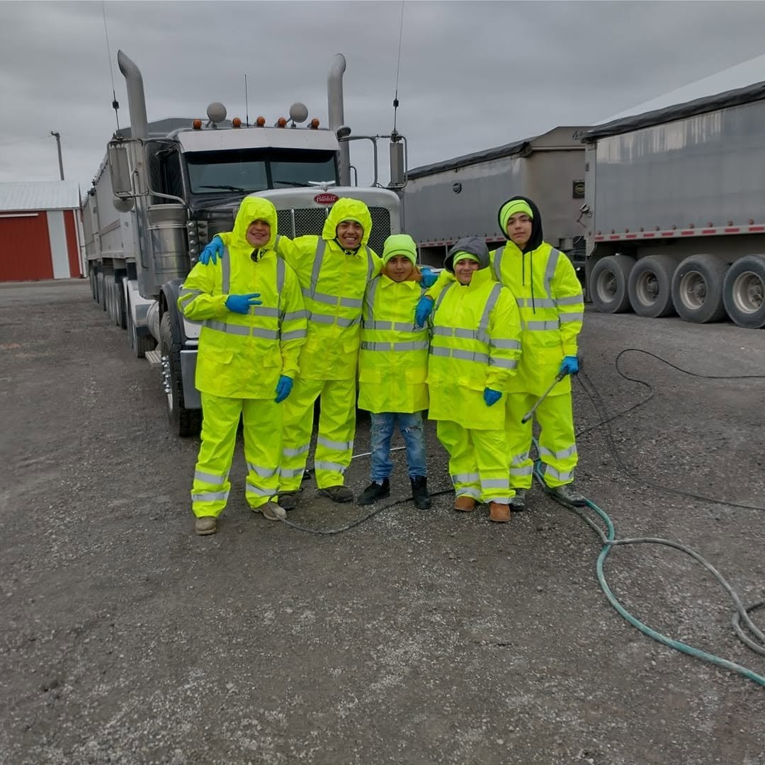 Five people in bright yellow-green rain suits and blue gloves standing arm in arm on a gravel lot by a dump-truck tractor.