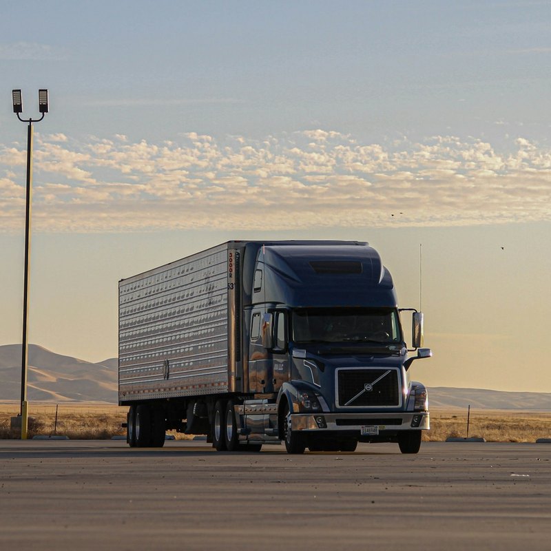 Blue tractor and polished ribbed aluminum trailer on a wide asphalt lot with dry hills and a pale sky behind.